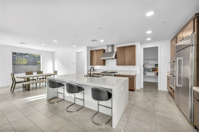 kitchen featuring sink, appliances with stainless steel finishes, an island with sink, a kitchen bar, and wall chimney exhaust hood