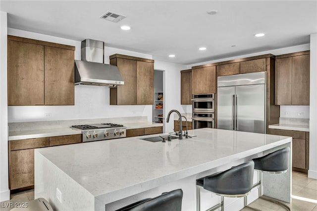 kitchen featuring wall chimney range hood, sink, a kitchen breakfast bar, stainless steel appliances, and a center island with sink