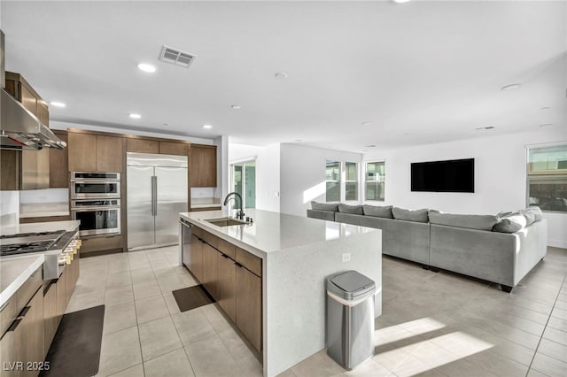 kitchen featuring light tile patterned flooring, appliances with stainless steel finishes, range hood, an island with sink, and sink