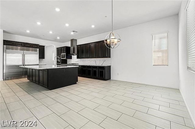 kitchen with hanging light fixtures, wall chimney range hood, tasteful backsplash, an island with sink, and appliances with stainless steel finishes