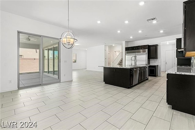 kitchen with light stone countertops, an island with sink, decorative light fixtures, ceiling fan with notable chandelier, and appliances with stainless steel finishes