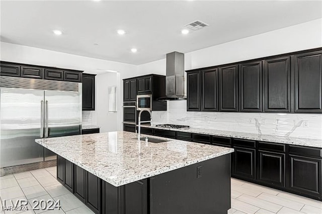 kitchen with light stone countertops, wall chimney exhaust hood, a kitchen island with sink, sink, and built in appliances