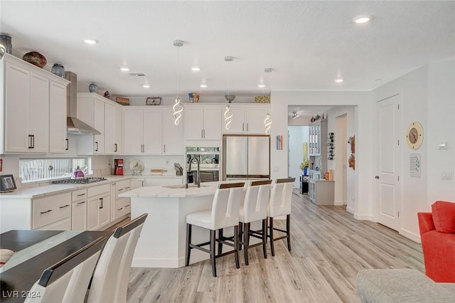 kitchen featuring wall chimney exhaust hood, hanging light fixtures, stainless steel appliances, a center island with sink, and white cabinets