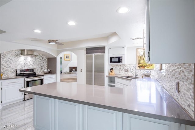 kitchen with built in appliances, a peninsula, a sink, white cabinets, and wall chimney range hood