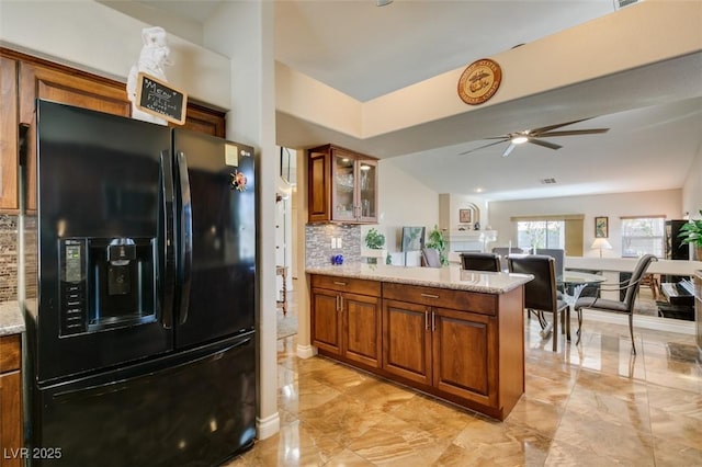 kitchen featuring ceiling fan, light stone counters, tasteful backsplash, black fridge with ice dispenser, and kitchen peninsula