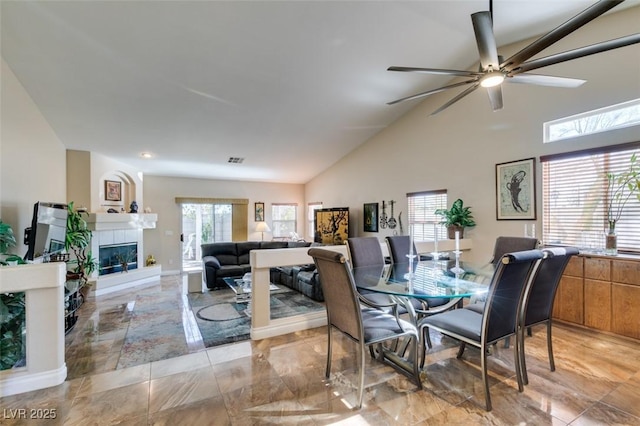 dining area featuring ceiling fan, a healthy amount of sunlight, high vaulted ceiling, and a tiled fireplace