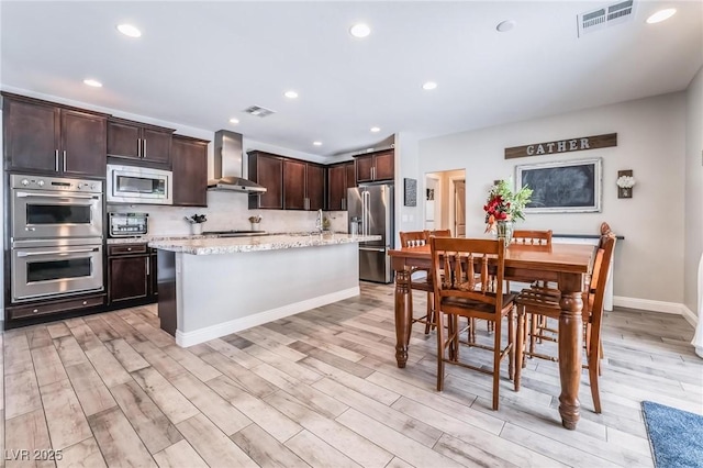 kitchen featuring wall chimney exhaust hood, light stone counters, light hardwood / wood-style floors, a kitchen island, and appliances with stainless steel finishes