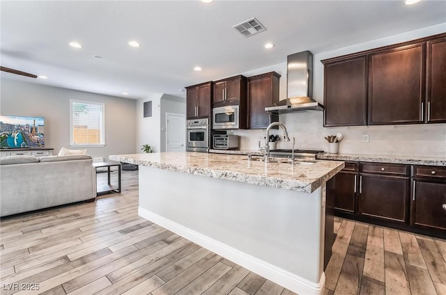 kitchen featuring a kitchen island with sink, wall chimney exhaust hood, stainless steel appliances, and light wood-type flooring