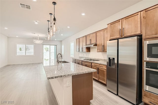 kitchen with appliances with stainless steel finishes, light stone counters, a kitchen island with sink, sink, and decorative light fixtures