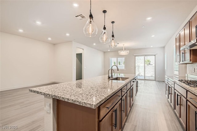 kitchen featuring sink, light wood-type flooring, an island with sink, decorative light fixtures, and light stone counters