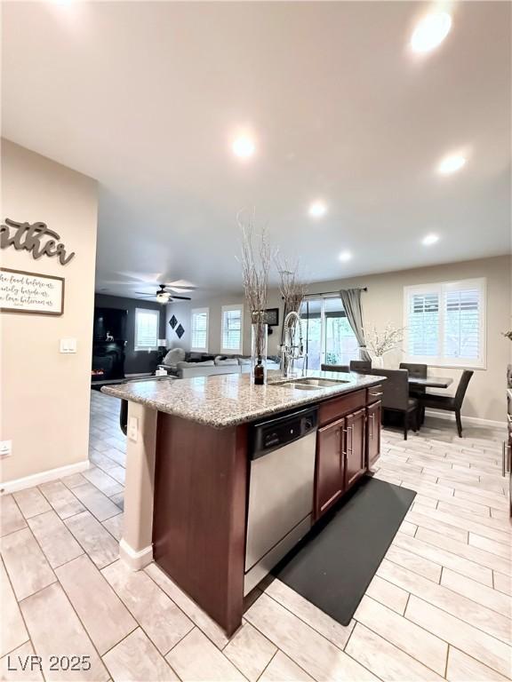 kitchen with sink, stainless steel dishwasher, an island with sink, dark brown cabinets, and light stone countertops