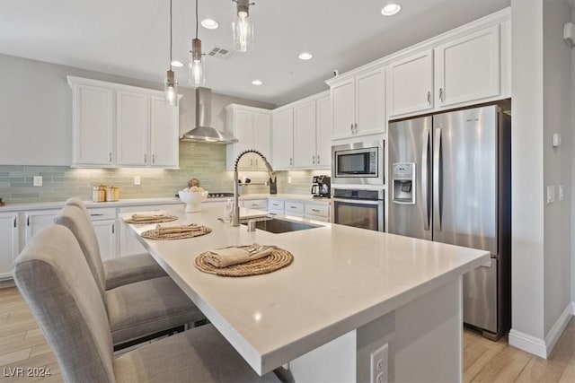 kitchen featuring white cabinets, wall chimney range hood, a kitchen island with sink, and appliances with stainless steel finishes