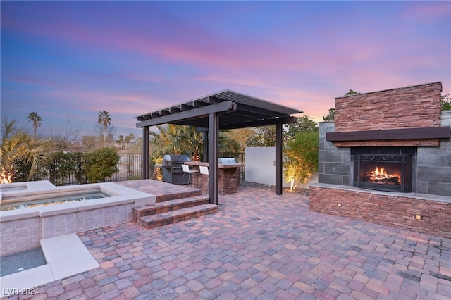 patio terrace at dusk with an outdoor kitchen, an outdoor stone fireplace, grilling area, and an outdoor hot tub