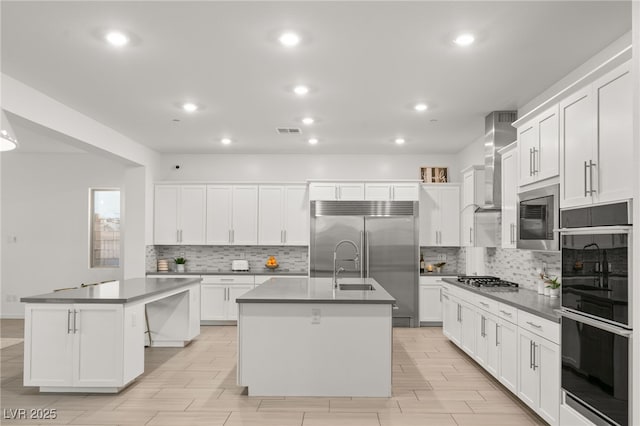 kitchen with white cabinets, wall chimney range hood, and an island with sink