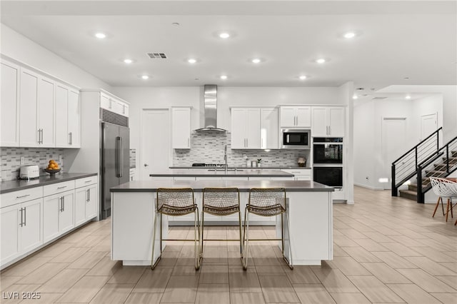 kitchen with built in appliances, wall chimney exhaust hood, a center island with sink, and white cabinetry