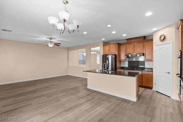 kitchen featuring a center island with sink, ceiling fan with notable chandelier, hanging light fixtures, light hardwood / wood-style floors, and stainless steel refrigerator