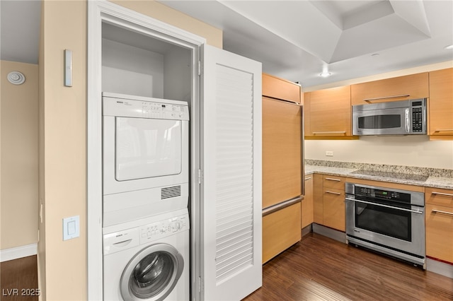 kitchen featuring stainless steel appliances, dark hardwood / wood-style floors, a tray ceiling, stacked washer / dryer, and light stone counters