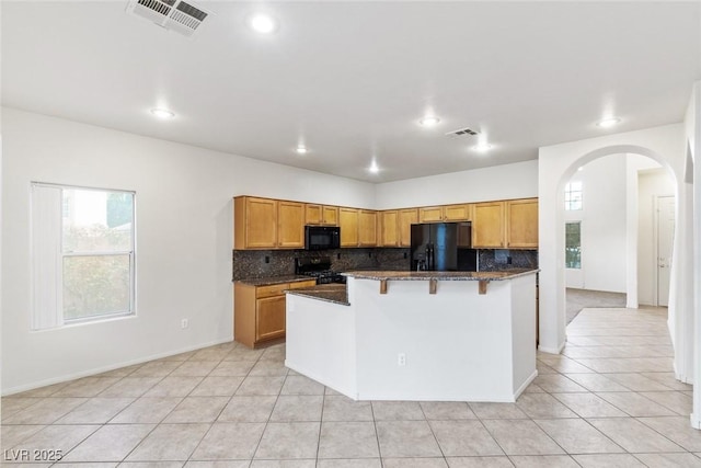 kitchen with light tile patterned floors, a center island with sink, dark stone counters, and black appliances