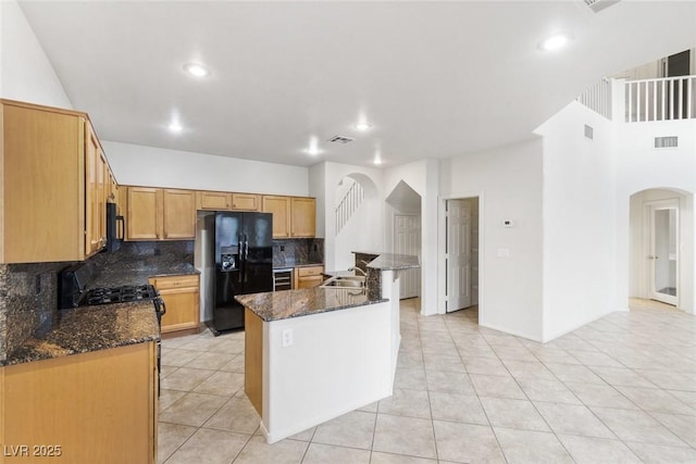 kitchen with decorative backsplash, sink, black appliances, dark stone countertops, and an island with sink