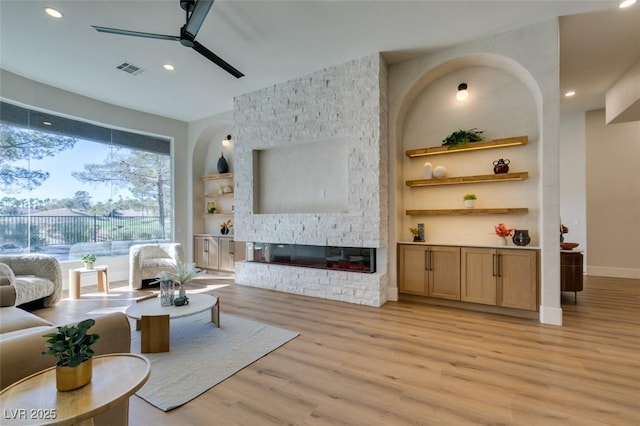 living room featuring built in features, light hardwood / wood-style flooring, a stone fireplace, and ceiling fan