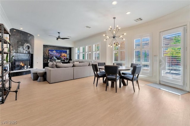 dining space with ceiling fan with notable chandelier, a tiled fireplace, a wealth of natural light, and light hardwood / wood-style flooring