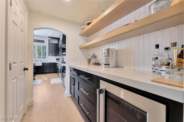 bar with light stone countertops, wall chimney exhaust hood, beverage cooler, light hardwood / wood-style flooring, and decorative backsplash