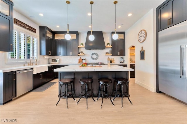 kitchen with sink, hanging light fixtures, stainless steel appliances, wall chimney range hood, and a kitchen island with sink