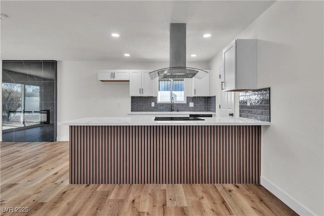 kitchen with kitchen peninsula, backsplash, island range hood, white cabinets, and light hardwood / wood-style floors