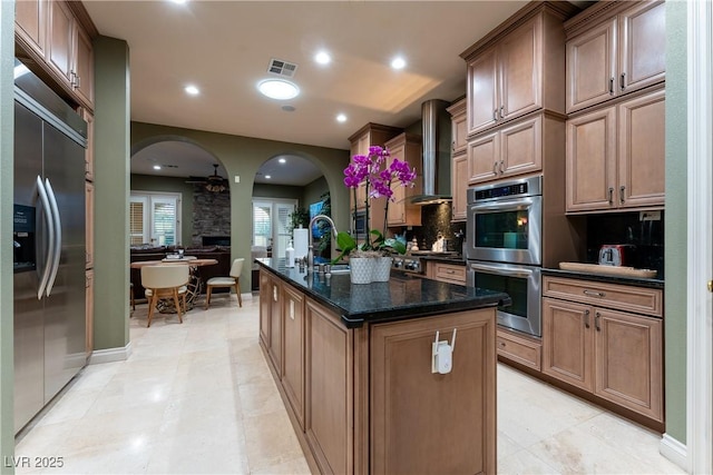 kitchen with sink, wall chimney exhaust hood, dark stone countertops, a kitchen island with sink, and appliances with stainless steel finishes