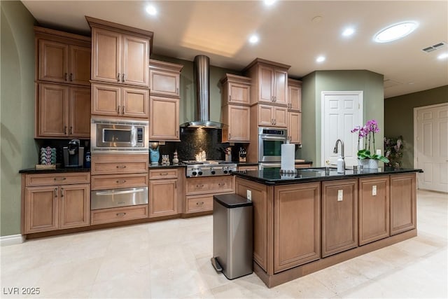 kitchen featuring appliances with stainless steel finishes, tasteful backsplash, dark stone counters, wall chimney exhaust hood, and an island with sink