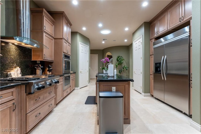 kitchen with tasteful backsplash, stainless steel appliances, a kitchen island with sink, wall chimney range hood, and dark stone countertops