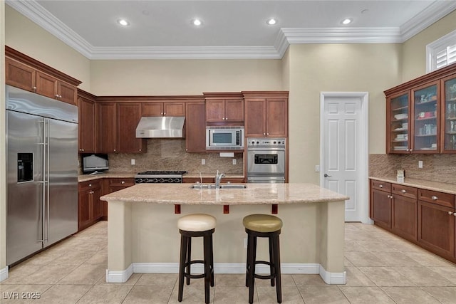 kitchen with a kitchen island with sink, sink, crown molding, built in appliances, and light stone counters