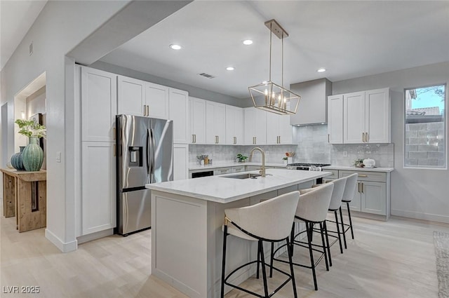 kitchen with white cabinetry, sink, wall chimney exhaust hood, stainless steel fridge with ice dispenser, and a center island with sink