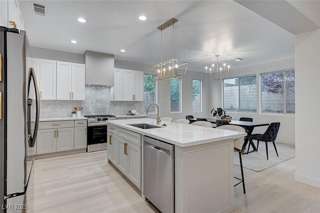 kitchen with sink, wall chimney range hood, a center island with sink, white cabinets, and appliances with stainless steel finishes