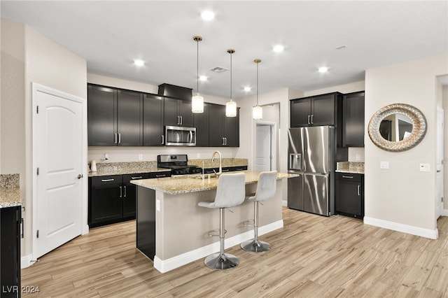 kitchen with light stone countertops, light wood-type flooring, stainless steel appliances, decorative light fixtures, and a center island with sink