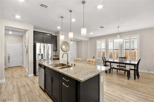 kitchen featuring a kitchen island with sink, sink, light stone countertops, decorative light fixtures, and stainless steel appliances