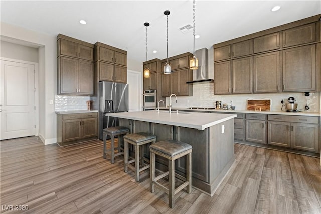 kitchen featuring a kitchen island with sink, light countertops, appliances with stainless steel finishes, wall chimney exhaust hood, and decorative light fixtures