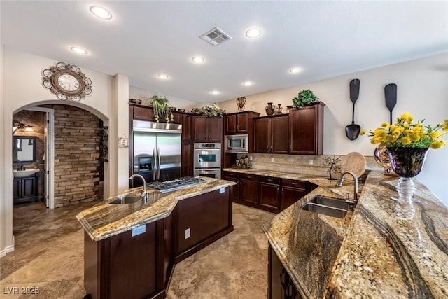 kitchen with light stone countertops, built in appliances, a kitchen island with sink, and sink