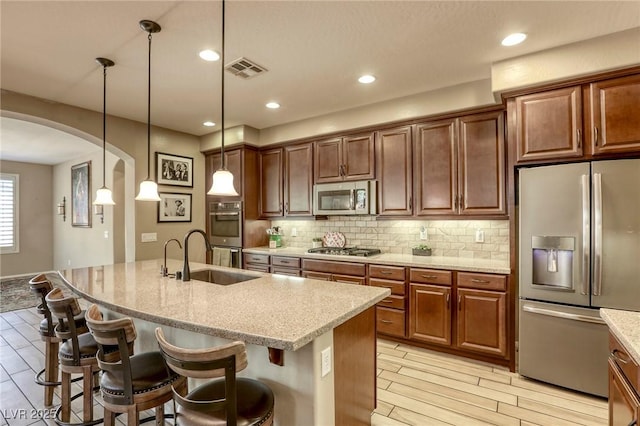 kitchen featuring light stone countertops, hanging light fixtures, tasteful backsplash, a center island with sink, and appliances with stainless steel finishes