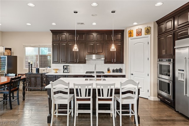 kitchen featuring decorative light fixtures, dark brown cabinets, dark hardwood / wood-style flooring, an island with sink, and stainless steel appliances