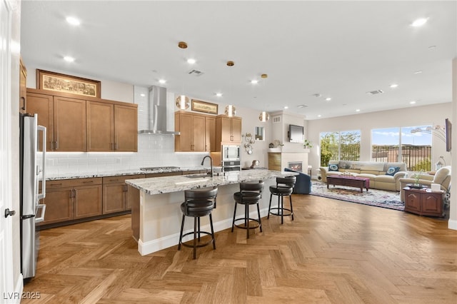 kitchen with a breakfast bar, light stone counters, a kitchen island with sink, and wall chimney exhaust hood