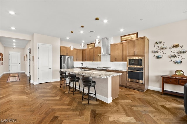 kitchen featuring a center island with sink, sink, wall chimney exhaust hood, light stone countertops, and stainless steel appliances