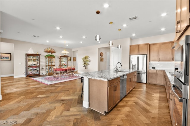 kitchen with sink, stainless steel appliances, light stone counters, decorative light fixtures, and a center island with sink