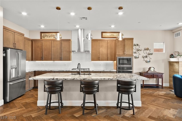 kitchen featuring wall chimney exhaust hood, stainless steel appliances, decorative light fixtures, and an island with sink