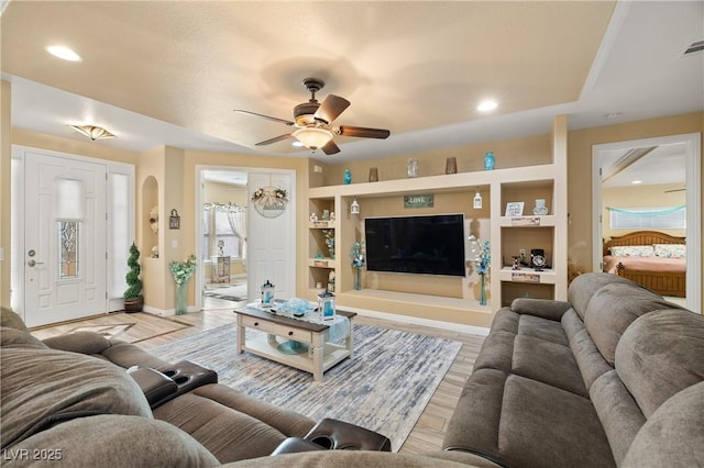 living room featuring built in shelves, ceiling fan, a raised ceiling, and light wood-type flooring