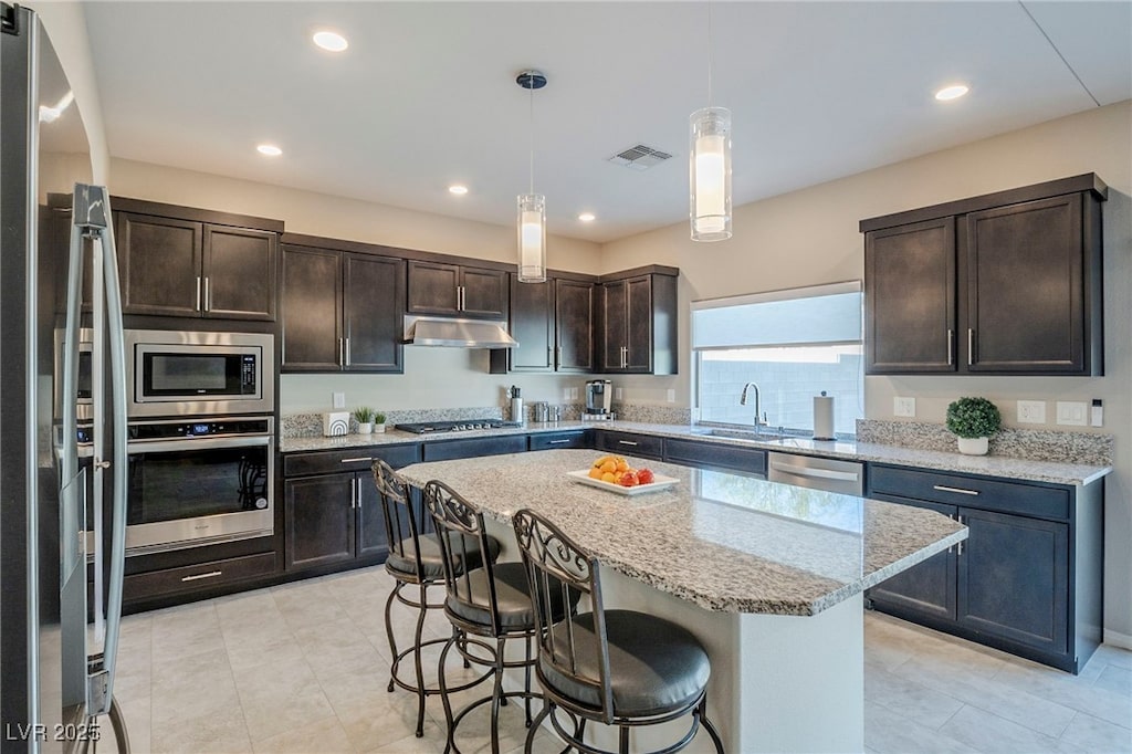 kitchen featuring hanging light fixtures, light stone counters, dark brown cabinets, a kitchen island, and appliances with stainless steel finishes