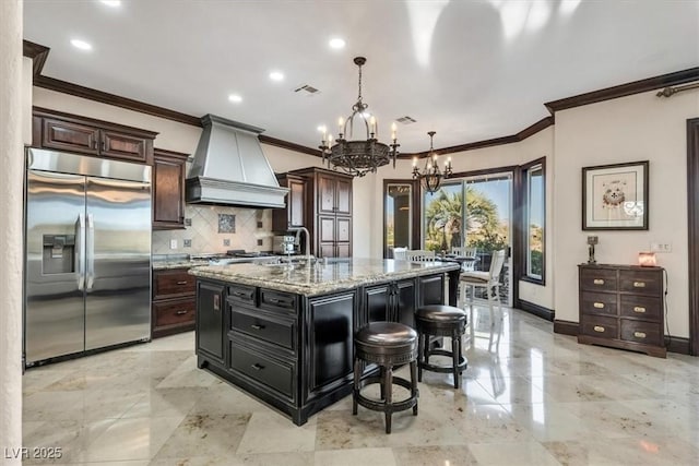 kitchen with light stone countertops, hanging light fixtures, built in refrigerator, a center island with sink, and custom exhaust hood