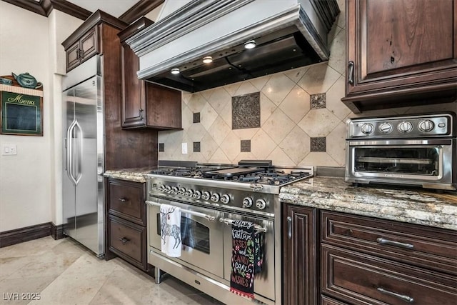 kitchen with high quality appliances, tasteful backsplash, dark brown cabinets, light stone counters, and custom range hood