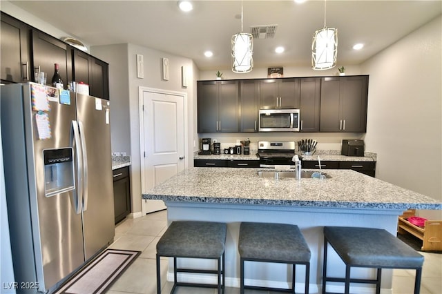 kitchen featuring appliances with stainless steel finishes, dark brown cabinets, decorative light fixtures, a center island with sink, and light tile patterned flooring