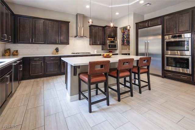 kitchen featuring hanging light fixtures, wall chimney exhaust hood, appliances with stainless steel finishes, a kitchen island, and a kitchen bar